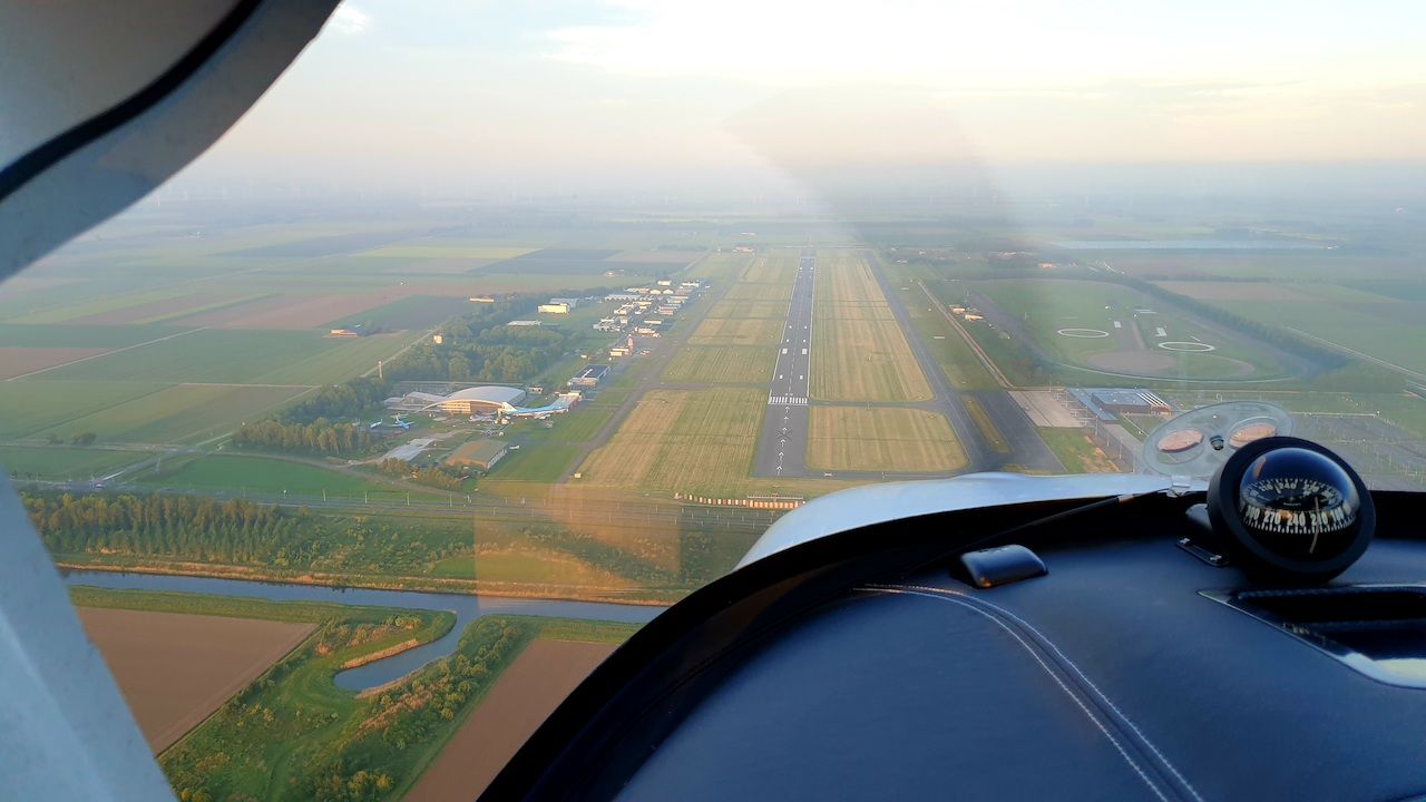 Uitzicht vanuit de cockpit op een naderende landingsbaan door de mist, met velden en luchthavengebouwen aan beide zijden.