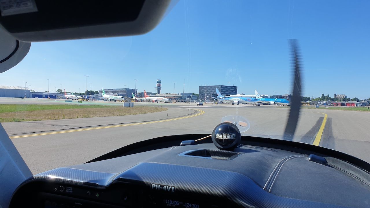 Uitzicht vanuit de cockpit op een taxibaan van het vliegveld, met de landingsbaan en terminal in de verte op een heldere dag.