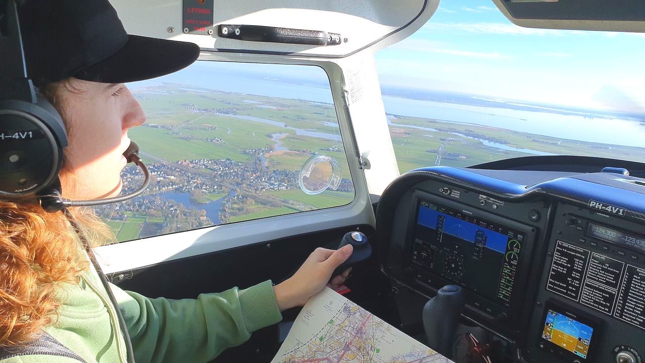 Een piloot zit in de cockpit en vliegt over een landschap, terwijl hij de stuurknuppel vasthoudt en een kaart op het dashboard raadpleegt.