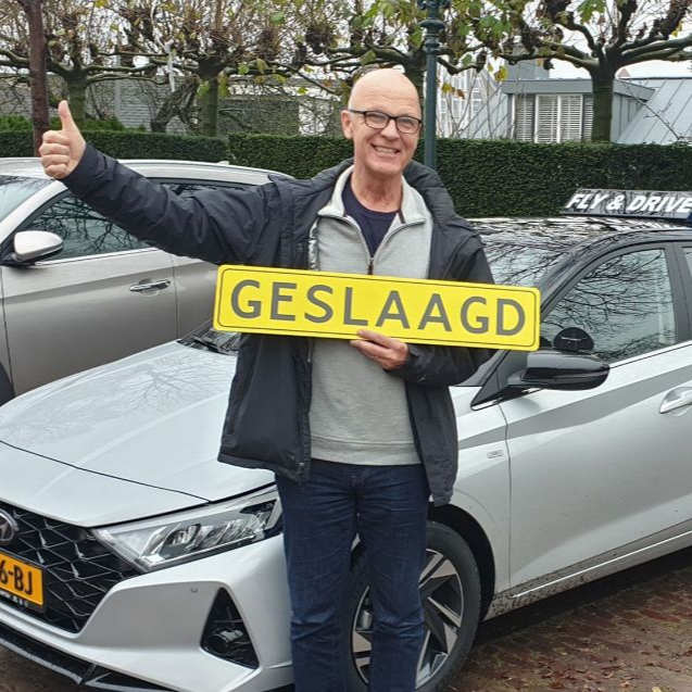 Een man staat lachend naast een zilverkleurige auto, houdt een geel