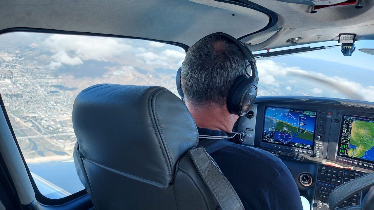 Een piloot bestuurt een klein vliegtuigje boven besneeuwde bergen, van achteren gezien, met de instrumenten in de cockpit zichtbaar.
