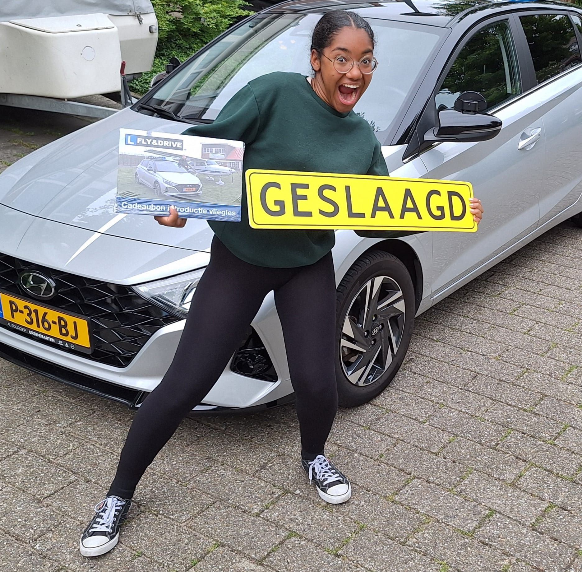 Een persoon poseert naast een zilverkleurige auto met een geel