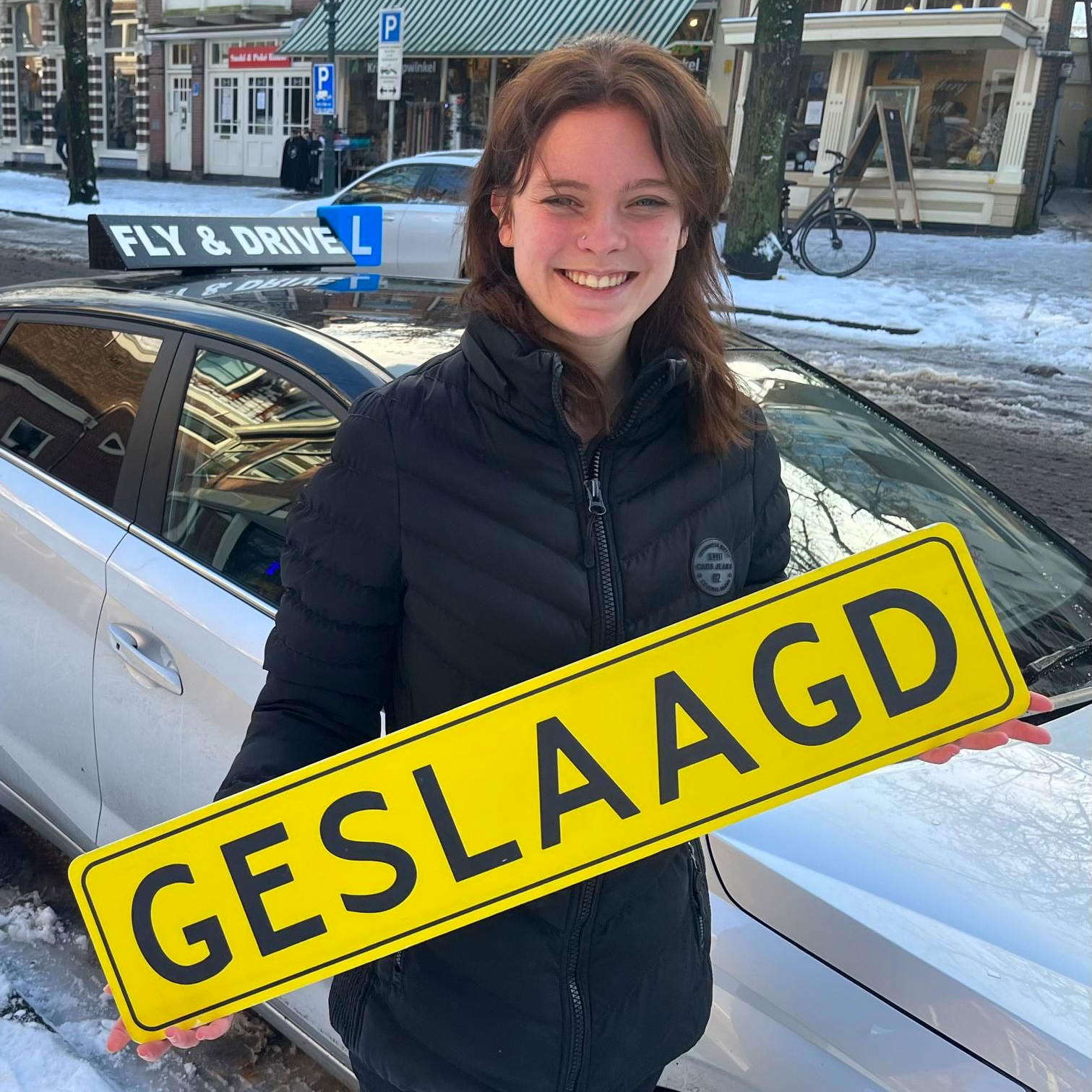 Een lachend persoon staat naast een witte auto, met op de voorgrond een groot geel