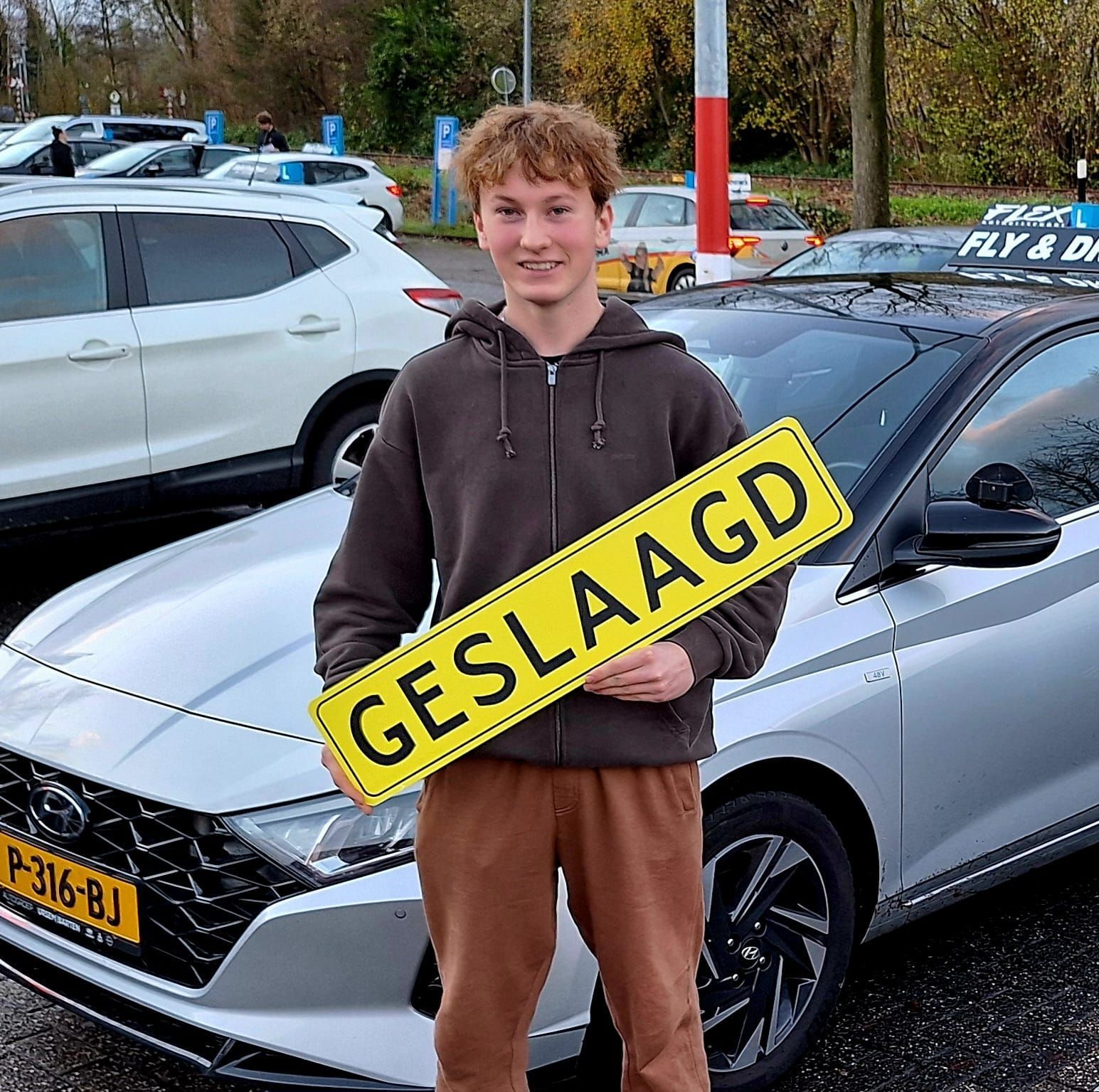 Een jonge man glimlacht naast een zilverkleurige auto en houdt een geel
