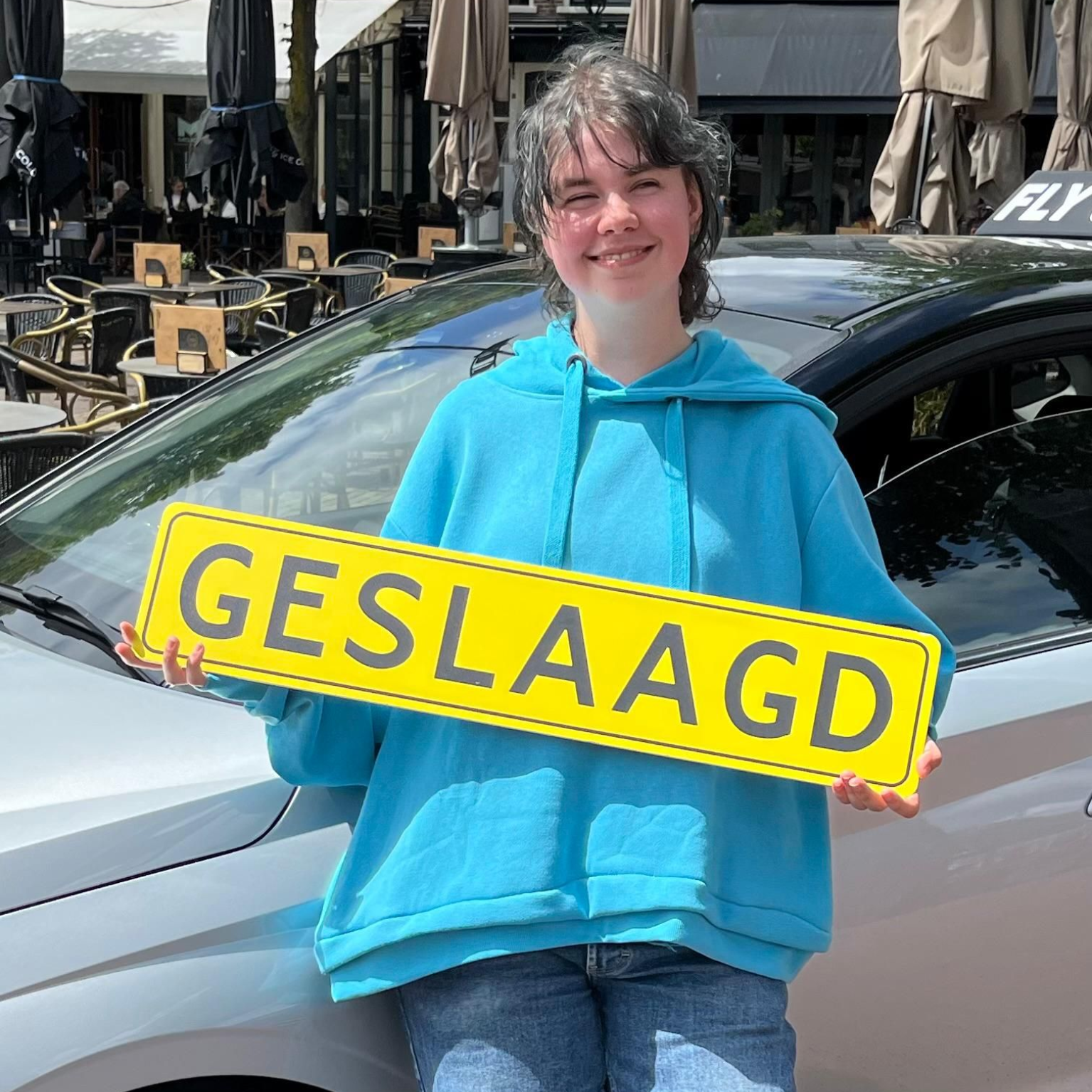 Een persoon in een blauwe hoodie lacht naast een auto en houdt een geel bord vast met de tekst