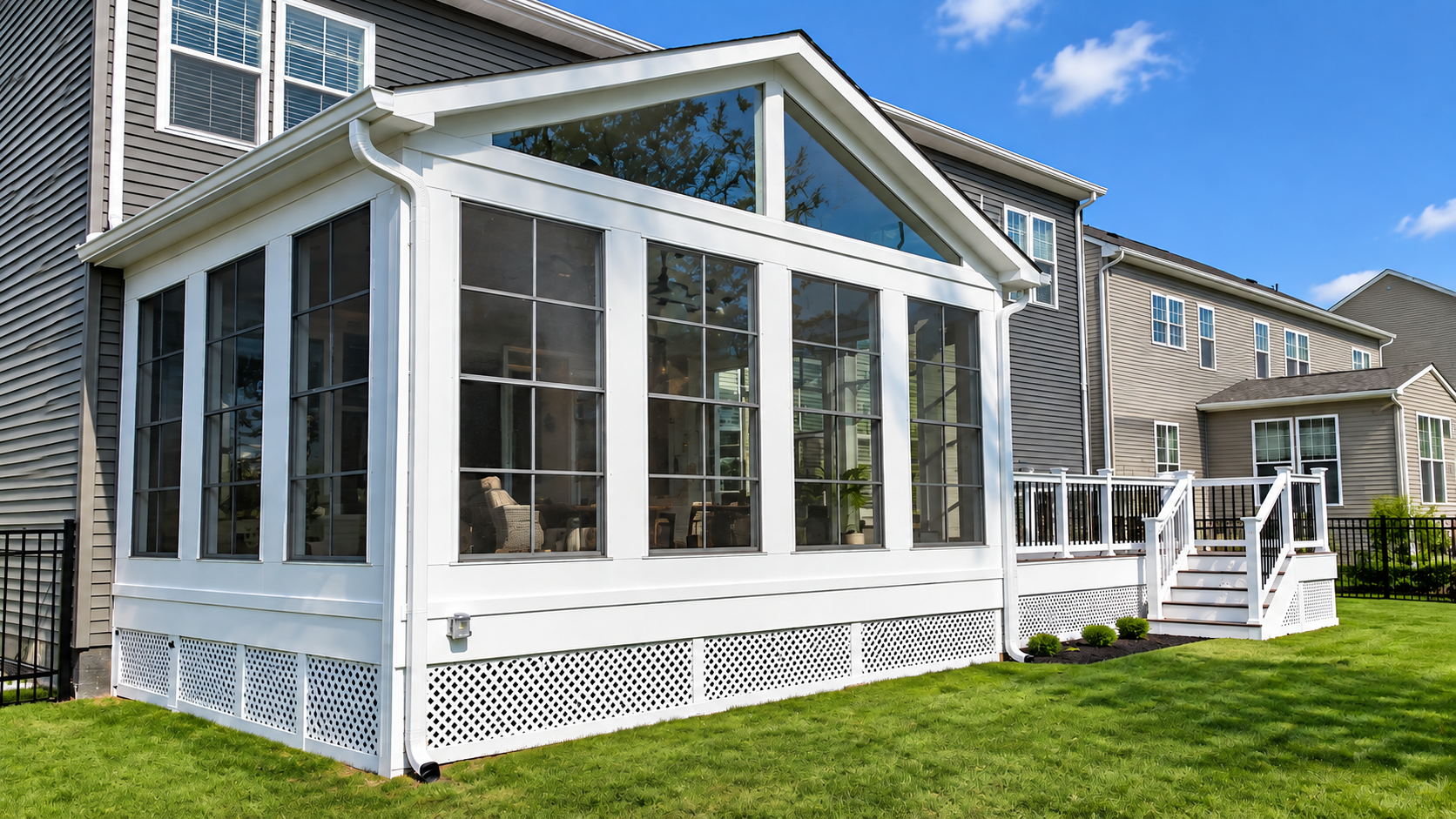 White sunroom addition with large windows and a lattice skirt beside a house on a green lawn