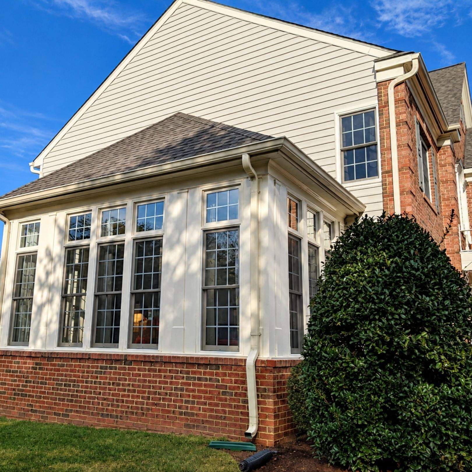 Brick house with large white-trimmed windows, beige siding, and a tall green shrub in front.