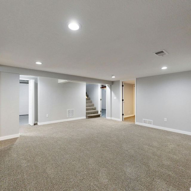 Empty carpeted basement room with white walls, recessed lights, and stairs in the back.