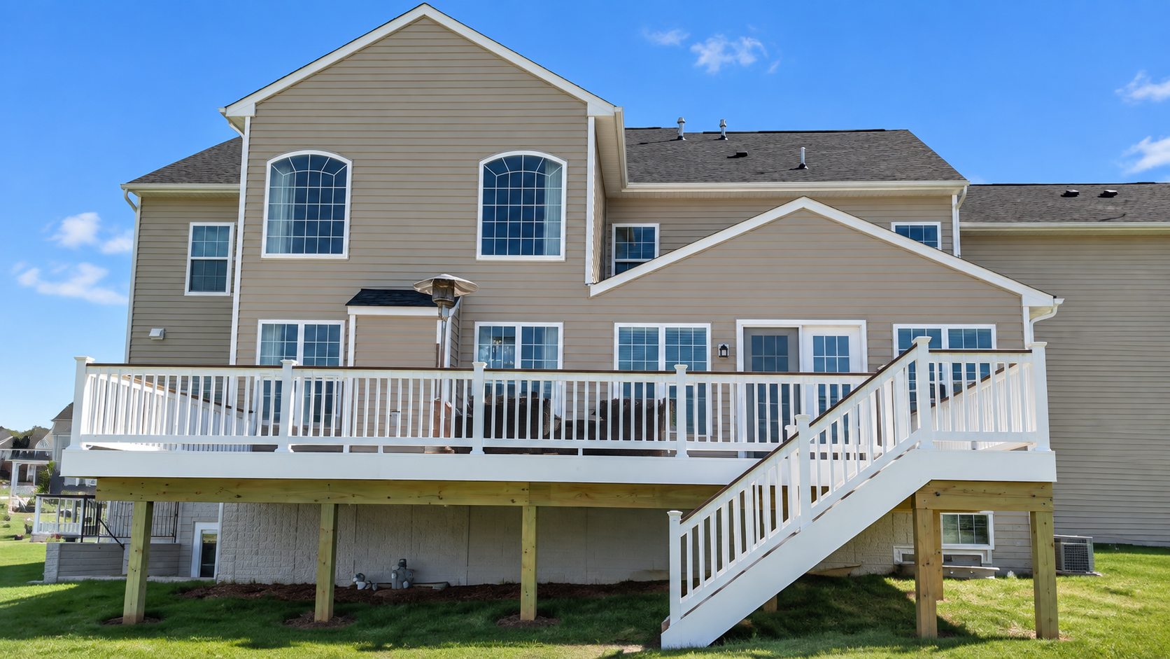 Two-story beige house with a white deck and stairs on a grassy yard under a blue sky