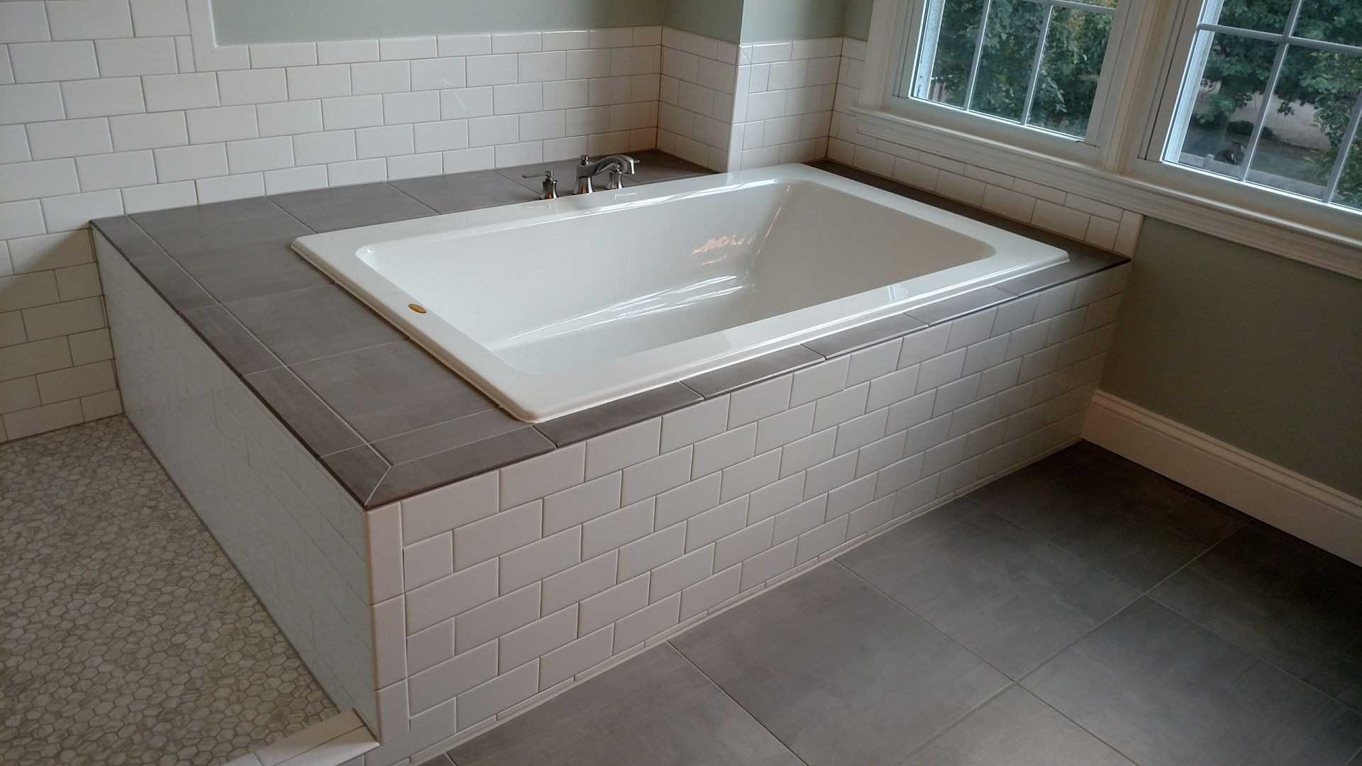 Modern bathroom with a rectangular built-in soaking tub, gray tile surround, and windows nearby