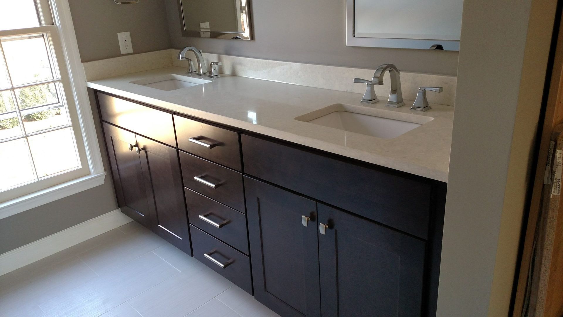 Bathroom vanity with dark cabinets, white countertop, double sinks, and a window on the left.