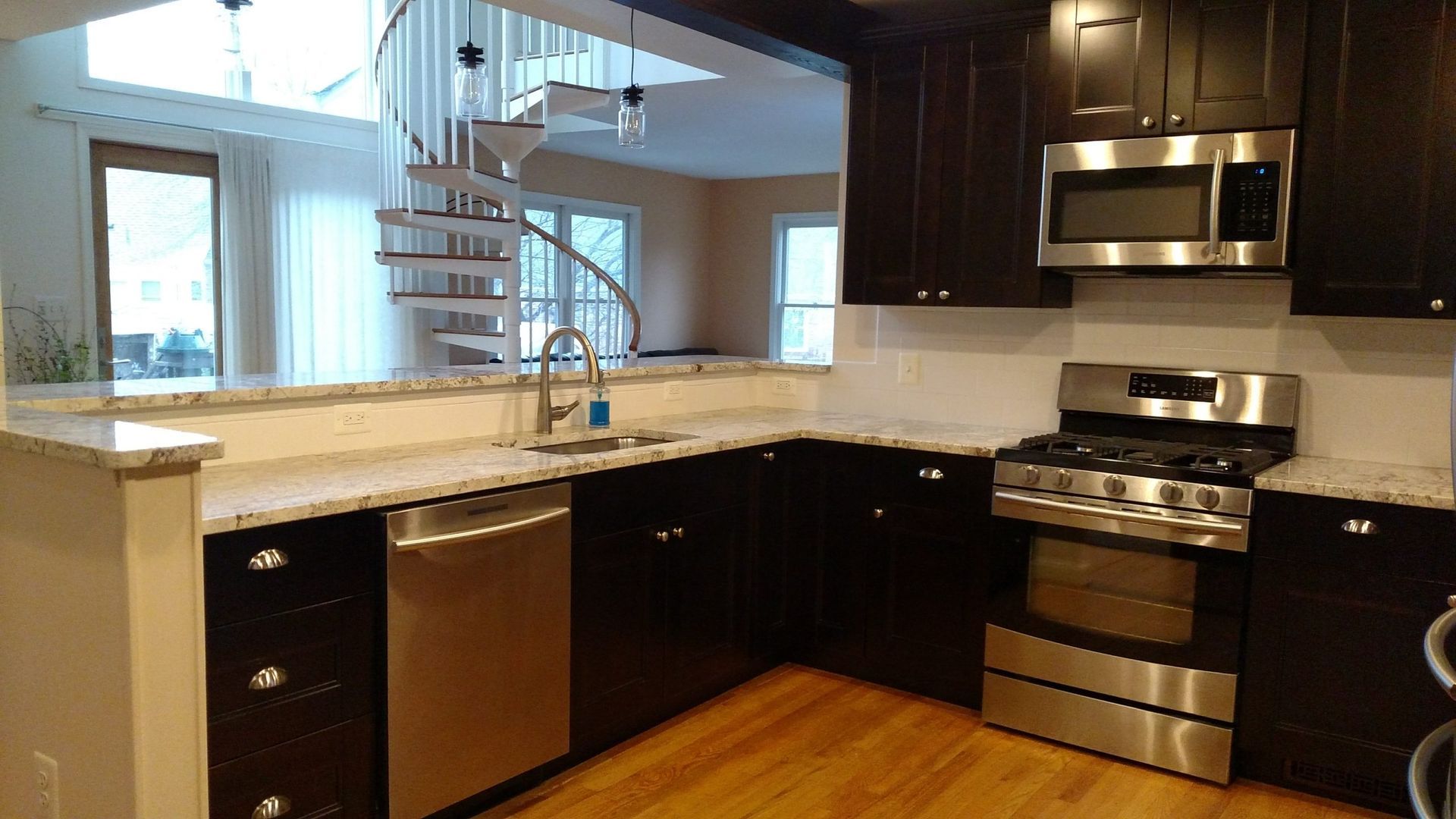 Modern kitchen with dark cabinets, stainless steel appliances, granite counters, and a spiral staircase by the window