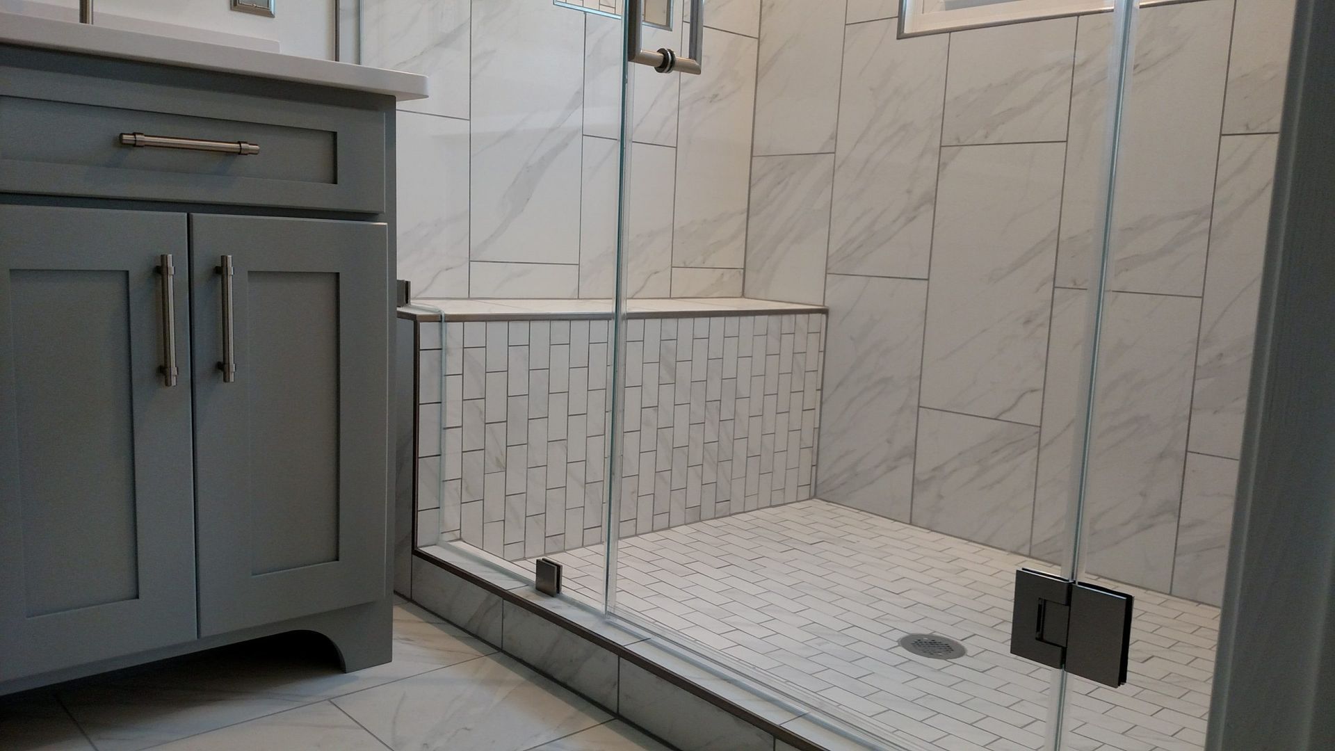 Modern bathroom with a gray vanity and glass shower enclosure over white marble tile.