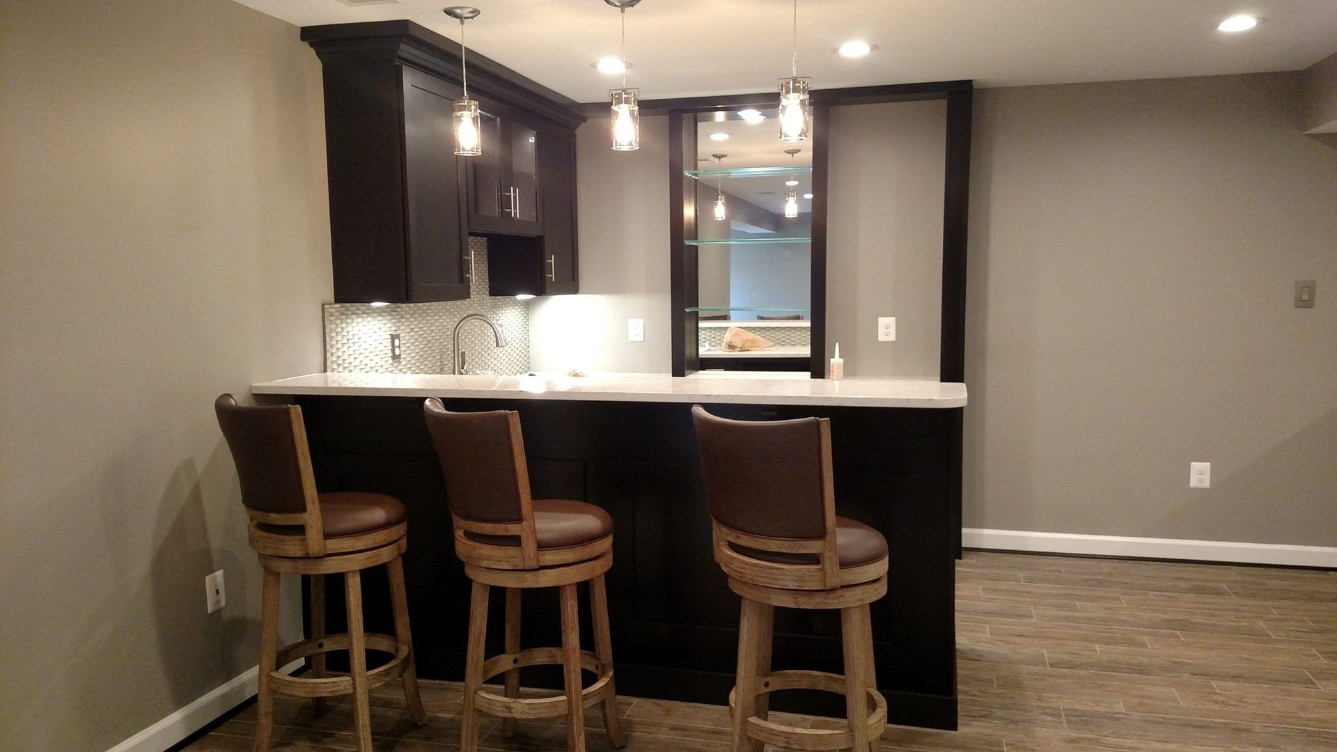 Empty home bar area with three wooden stools, granite counter, and open kitchen lighting