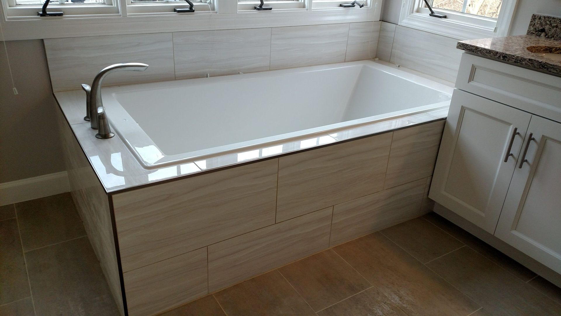 White freestanding bathtub by a window in a tiled bathroom, with chrome faucet and white cabinets