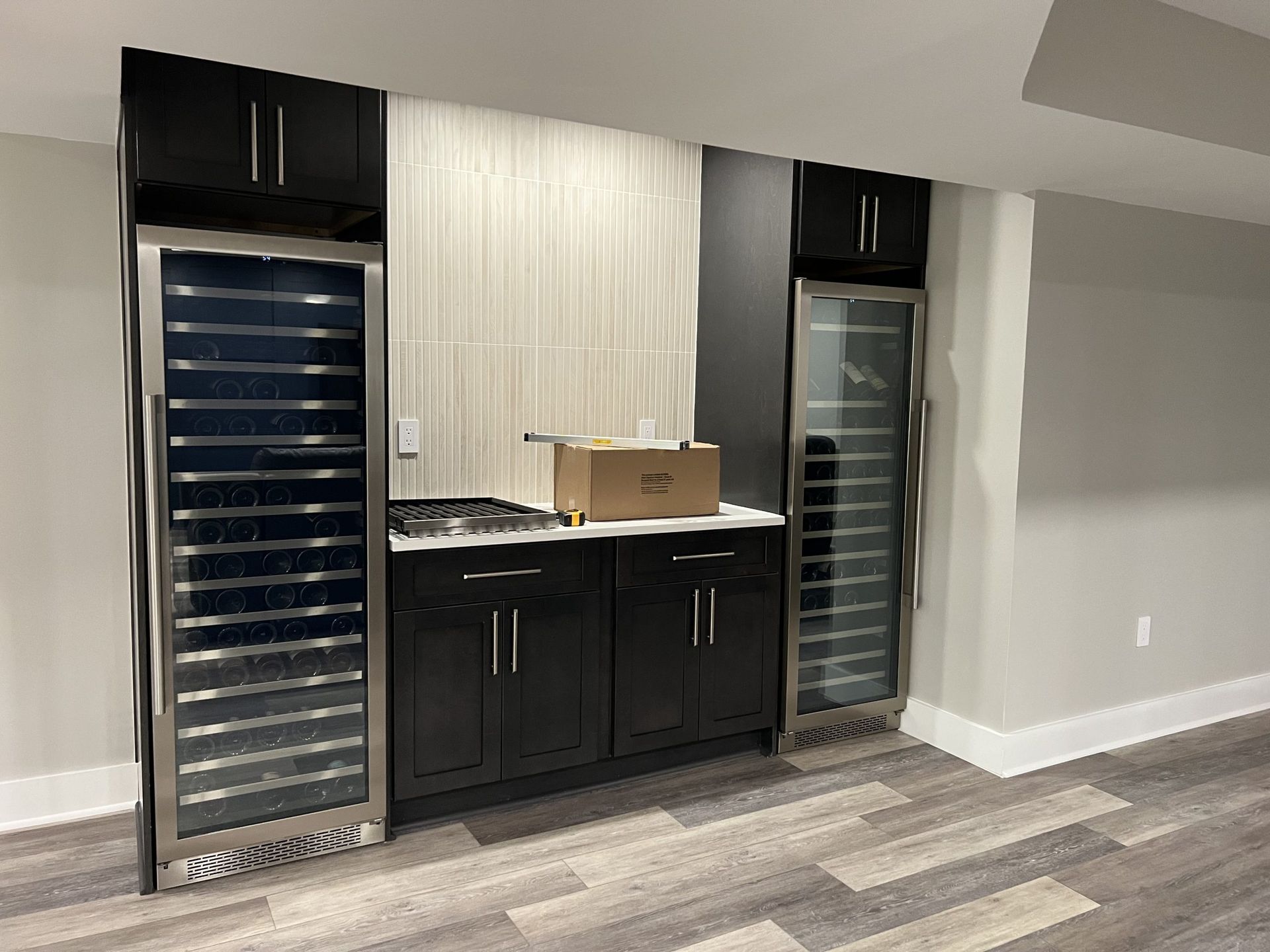 Modern wet bar with black cabinets, two glass-door wine fridges, and a cardboard box on the counter.