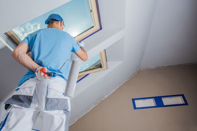 Painter on a step ladder painting a ceiling skylight, wearing blue shirt and cap in a room under renovation