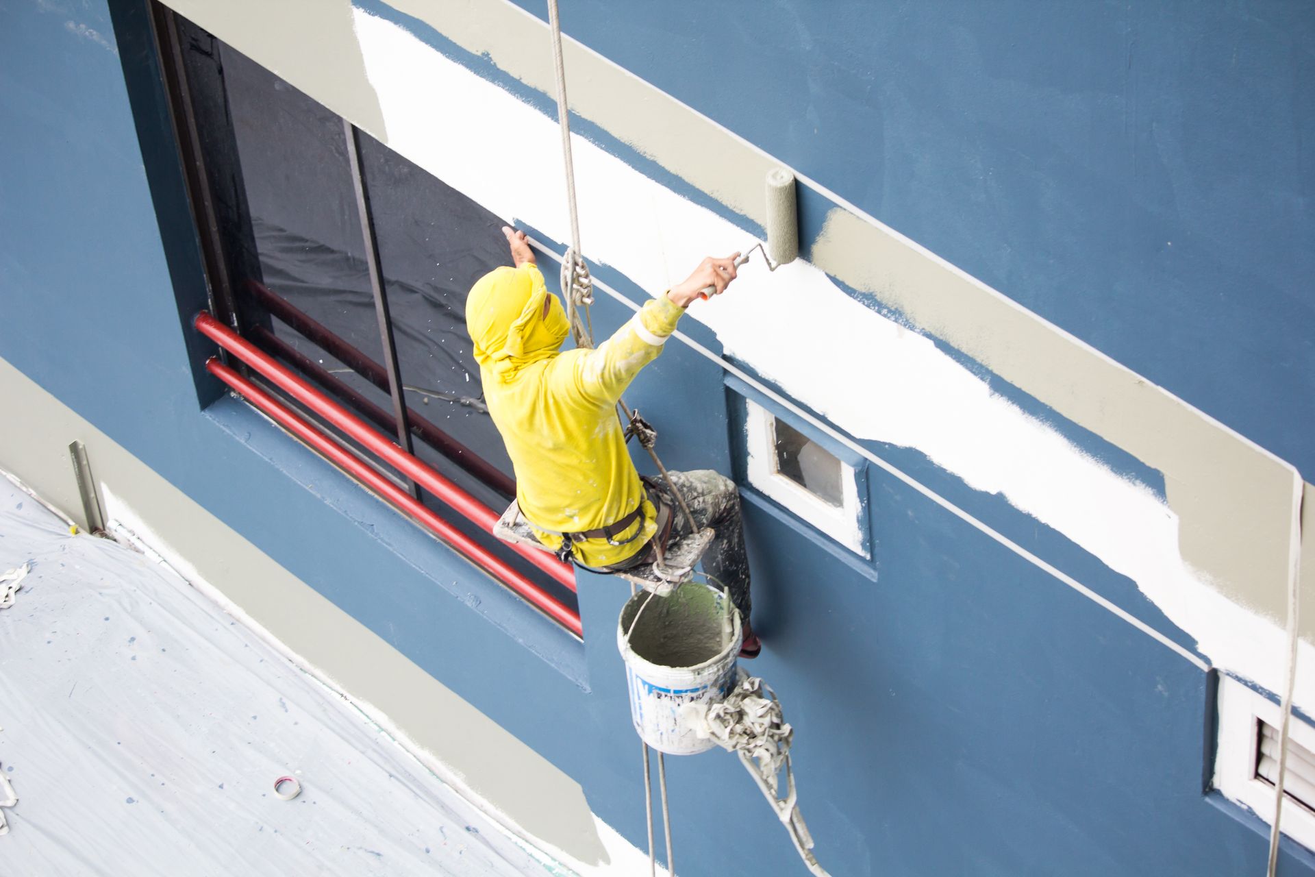 Worker in yellow hoodie painting a blue building from a bucket lift above a window