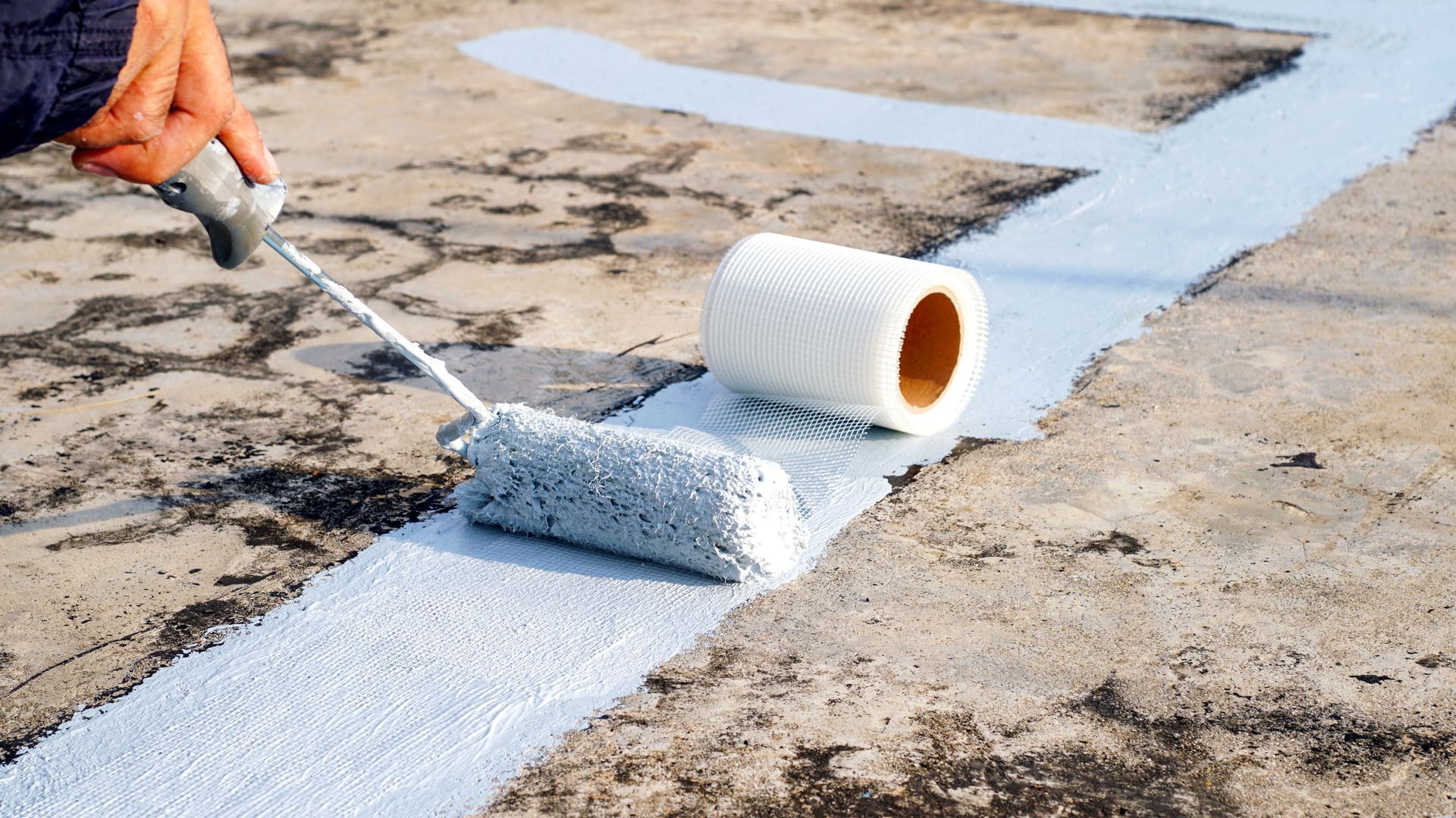 Hand using a paint roller to apply white coating on a cracked concrete surface