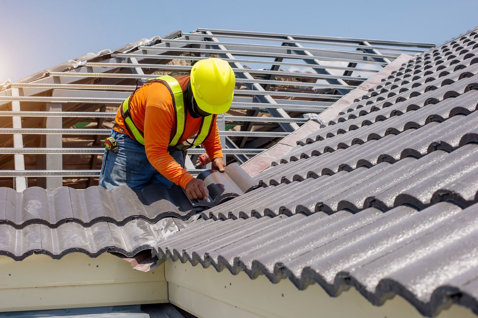 Roof worker in orange vest and yellow hard hat repairing gray tiled roof with power tool