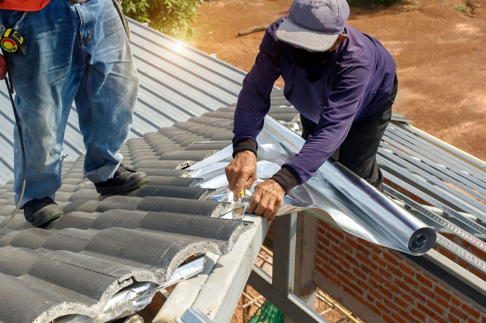 Two workers repairing a corrugated metal roof beside a brick wall, using tools and gloves