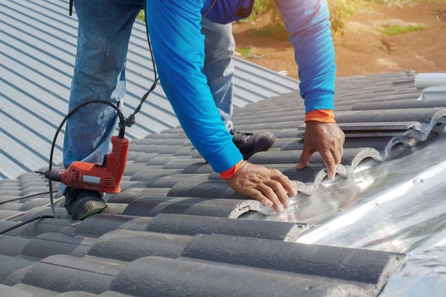 Worker repairing a gray tiled roof with a red power tool and flashing sheet metal