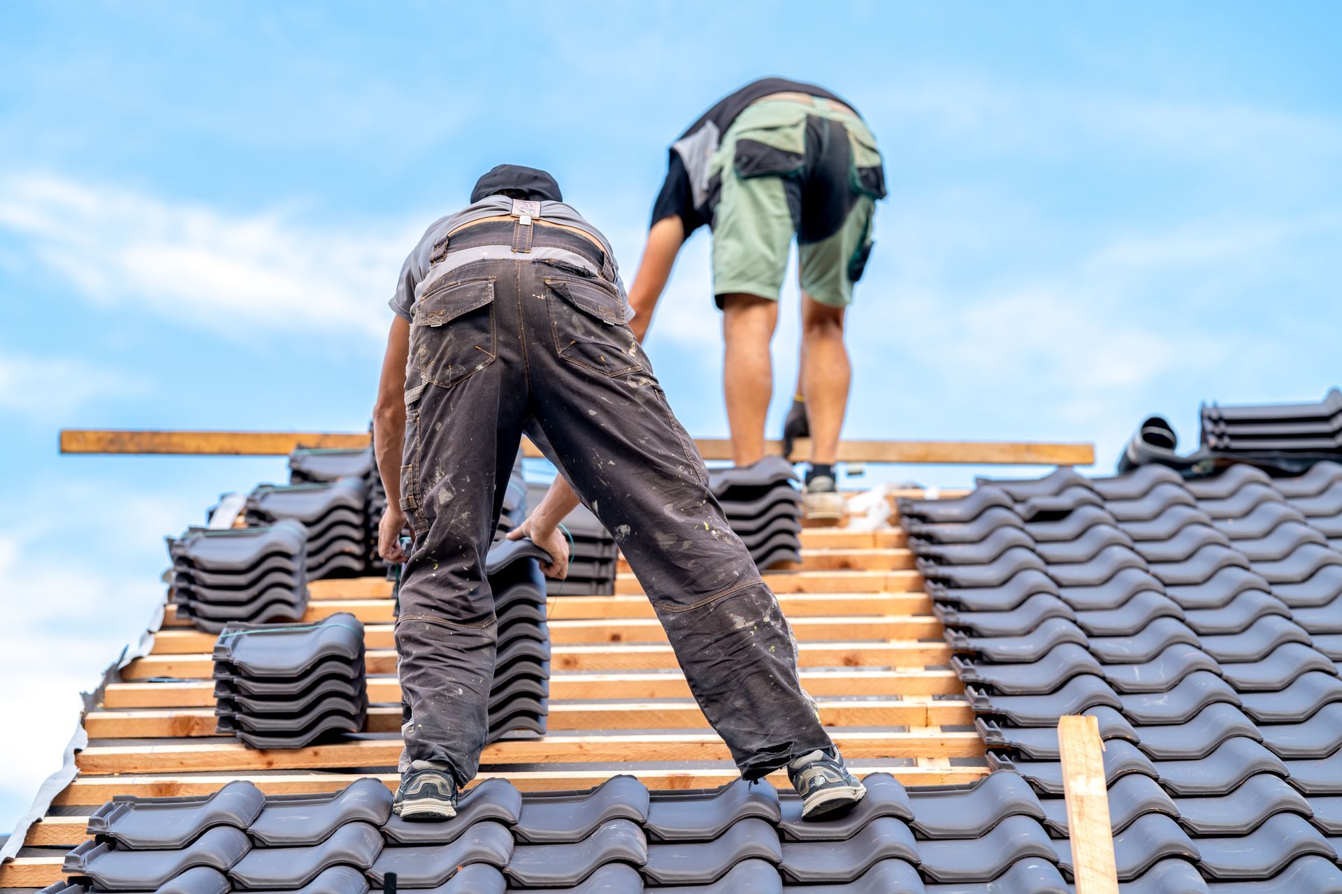 Two workers installing roof tiles on a house roof under a blue sky