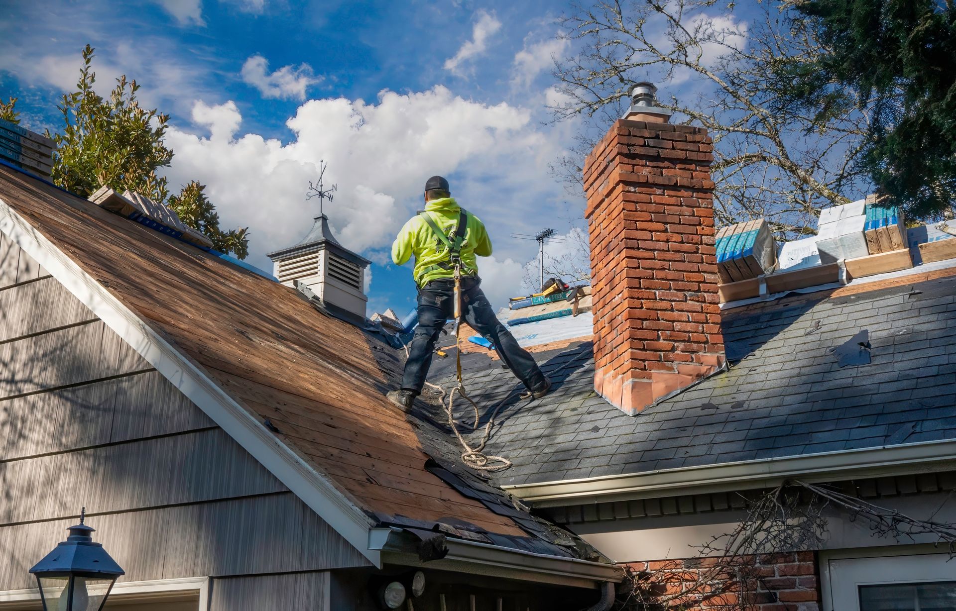 Roofing worker in a neon jacket repairing a shingled roof beside a brick chimney under a cloudy sky