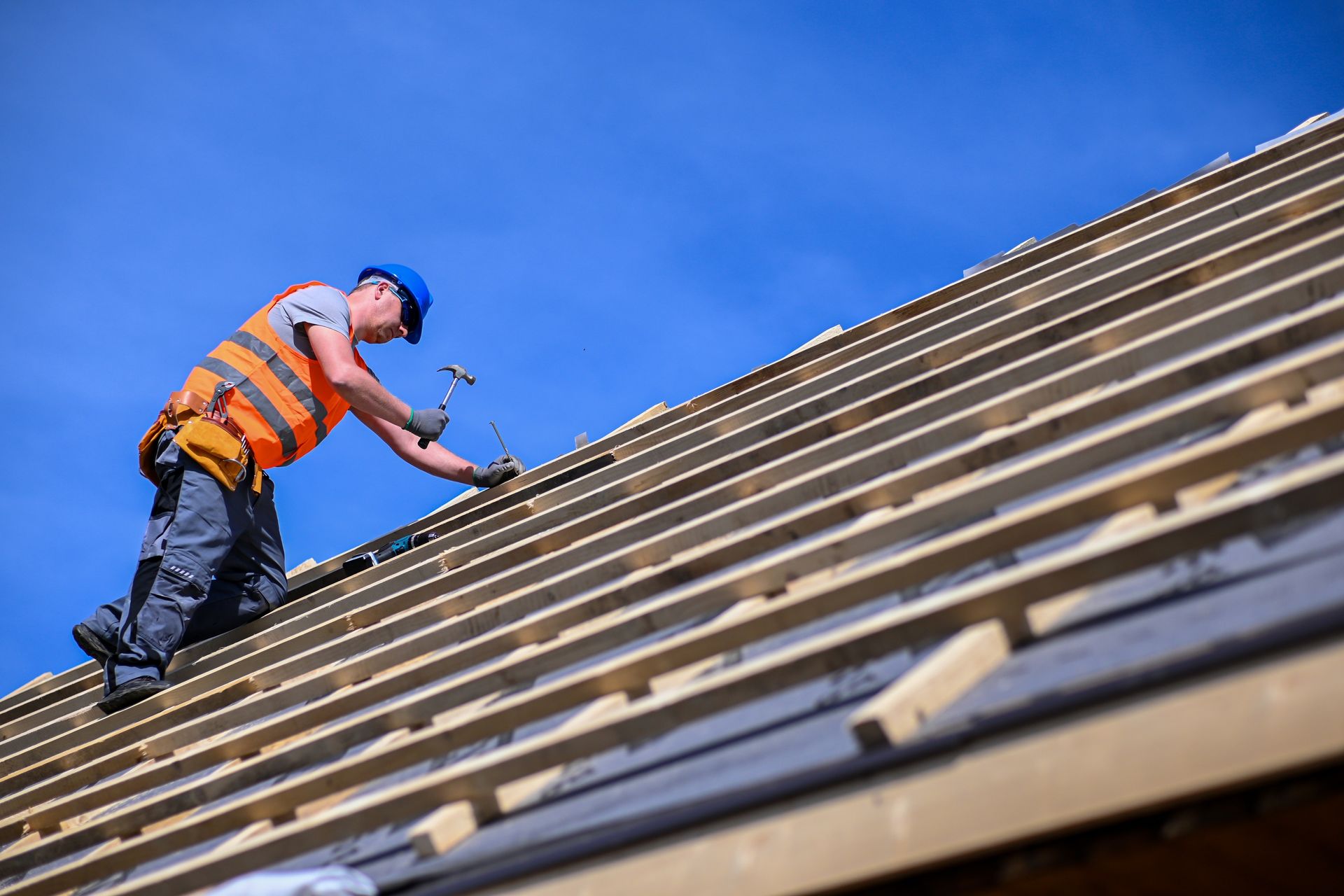 Worker in orange safety vest installing shingles on a steep roof under a blue sky