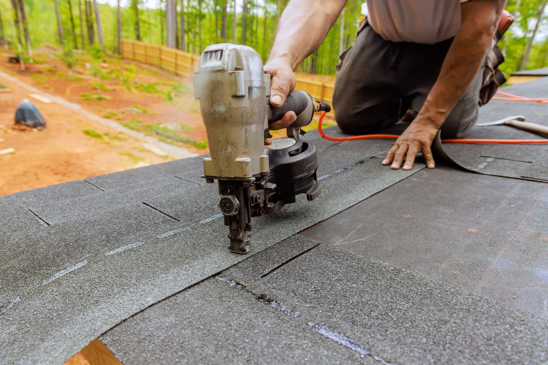 Worker using a nail gun to install black roofing shingles on a sloped roof