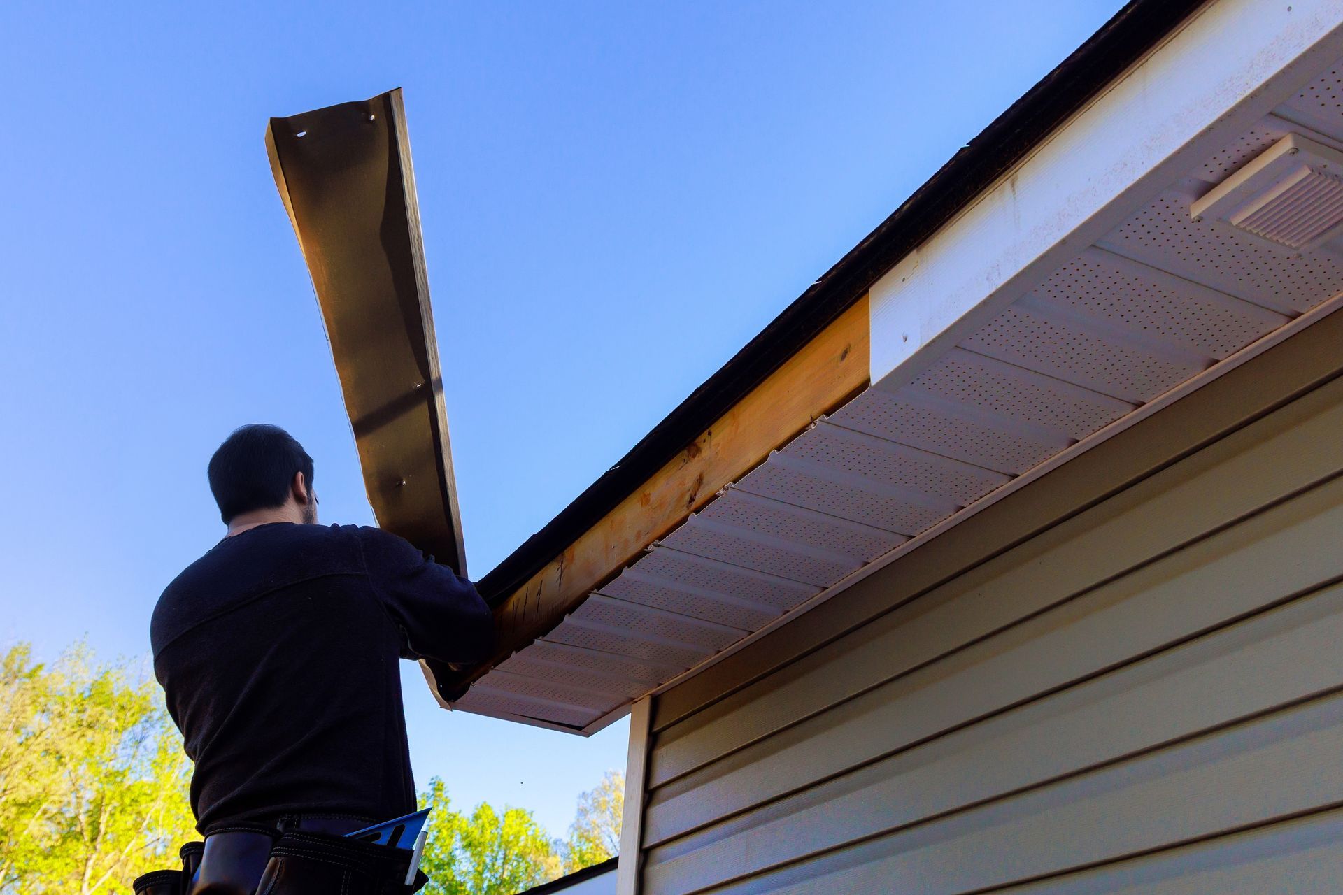 Person working on roof gutter beside a house, holding a long piece of siding or trim.