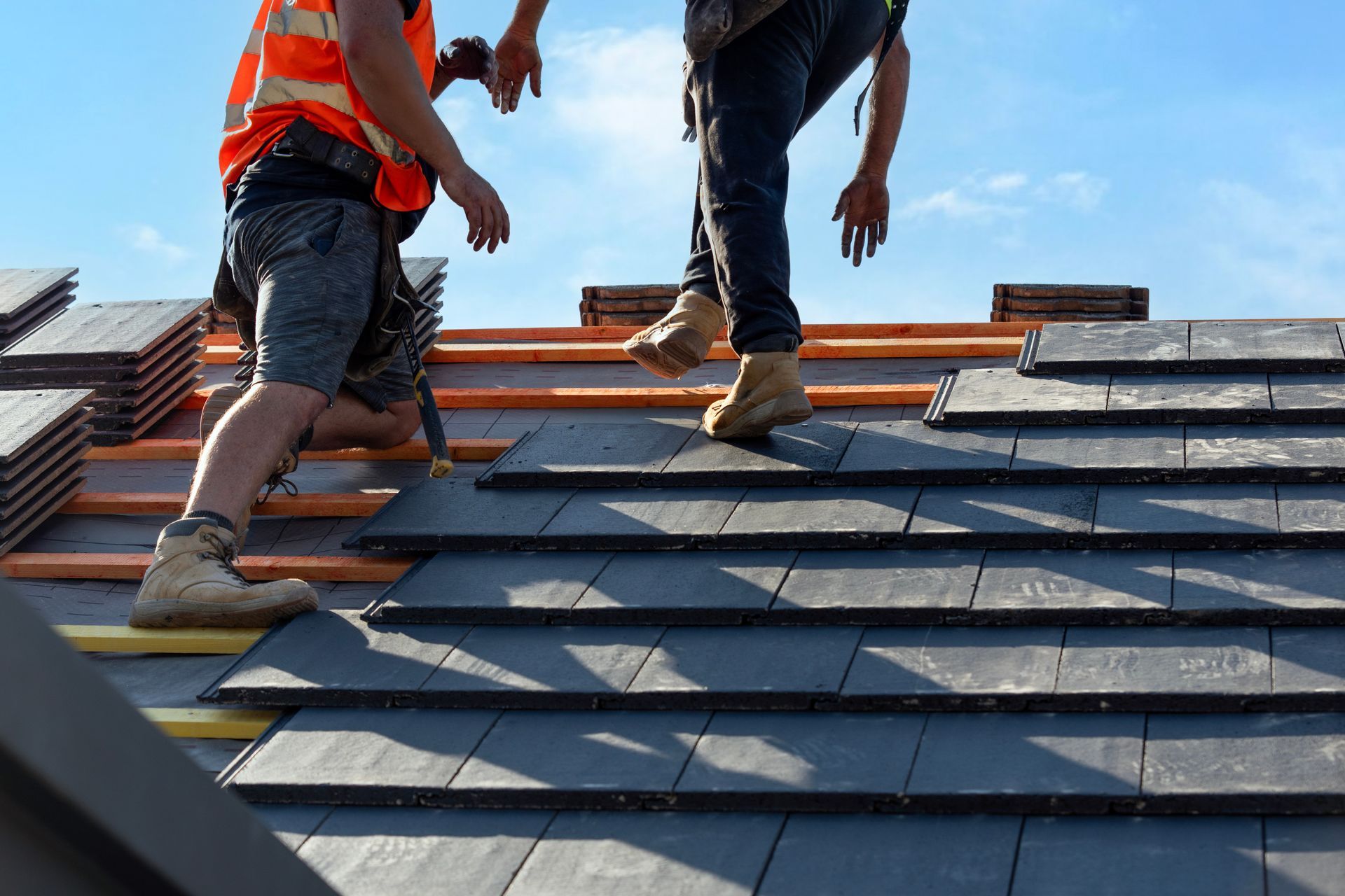 Workers installing gray roof shingles on a sloped roof under a blue sky