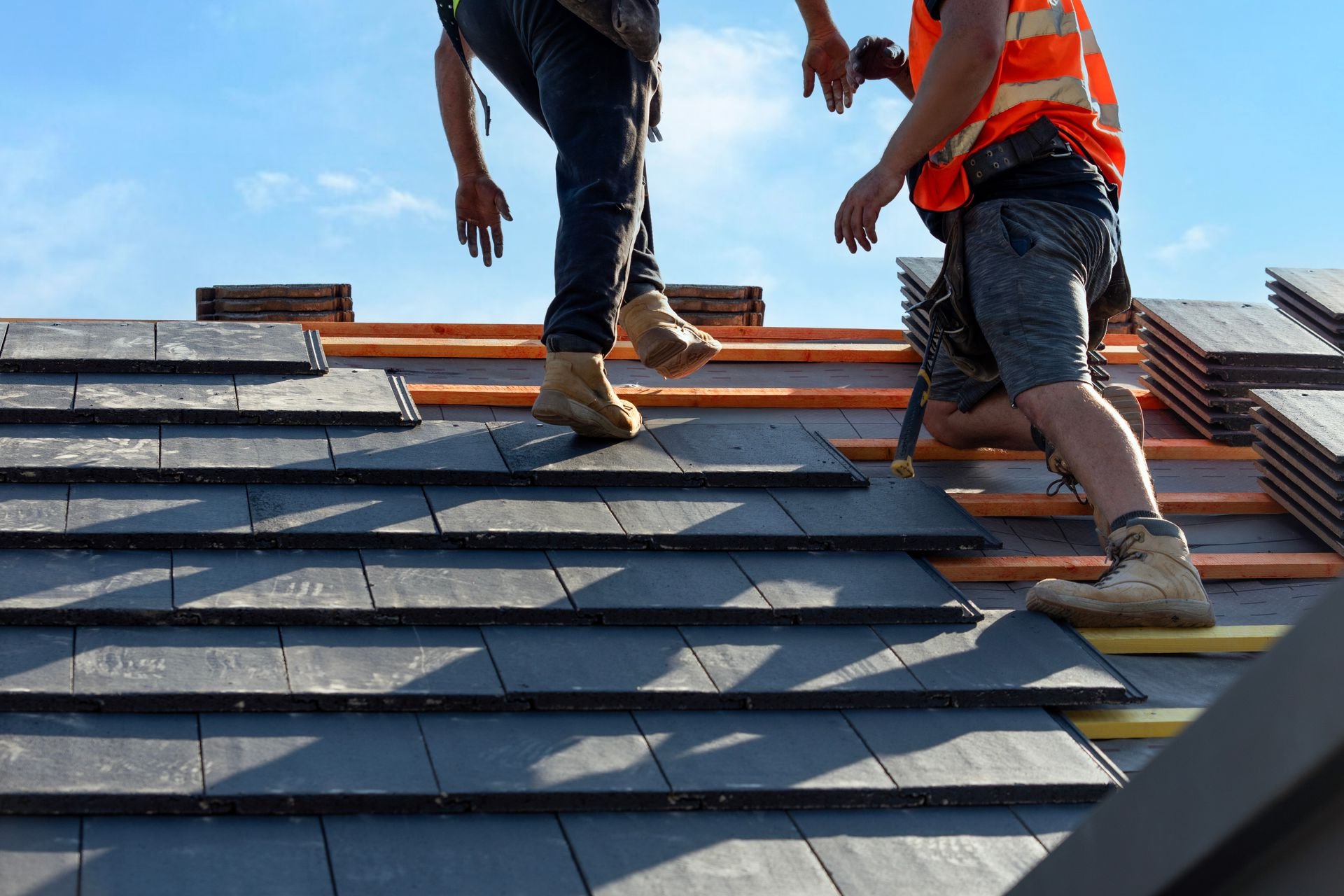 Two workers installing dark roof shingles on a sloped roof, one in an orange safety vest