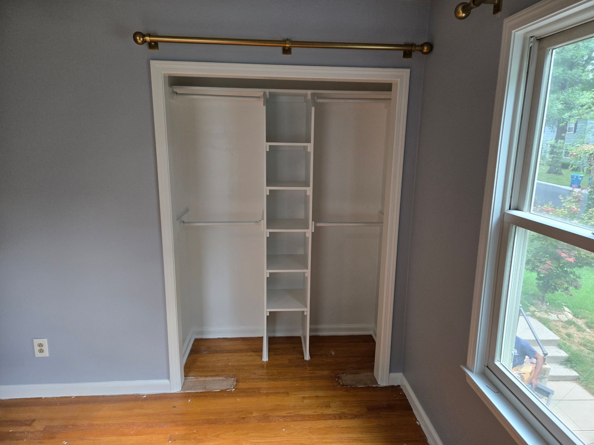 A bedroom closet with shelves, rods, and white trim. Wooden floor and a window to the right.