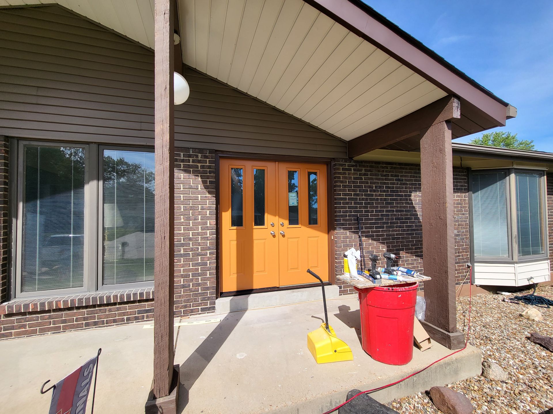 A red trash can is on the porch of a brick house.