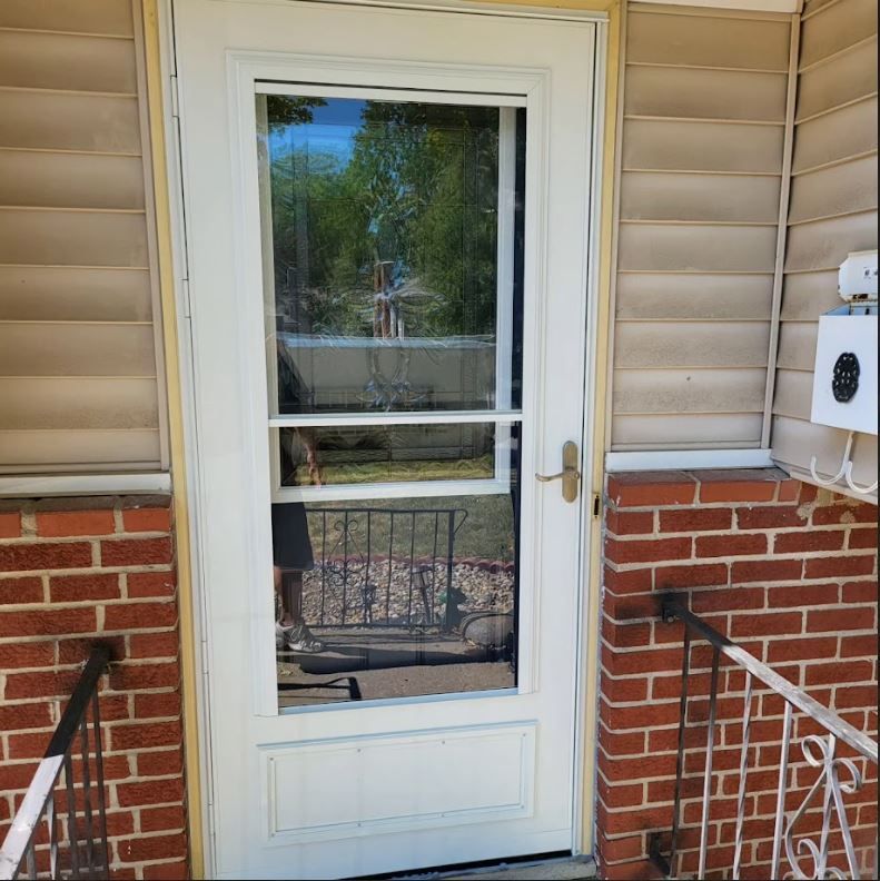 A white screen door is open on a brick house.
