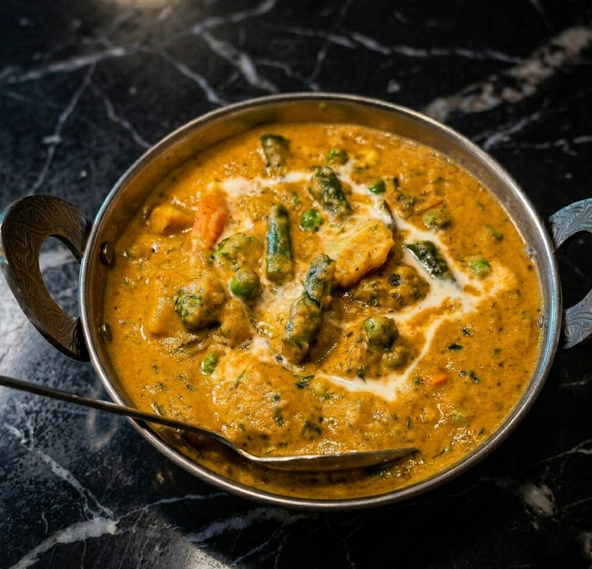 A silver bowl of vegetable curry garnished with cream, served on a black marble table with a spoon.