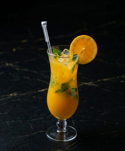 An orange-colored cocktail with mint and an orange slice garnish in a curved glass on a black marble surface.