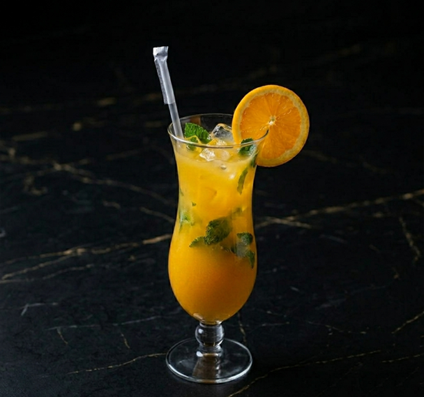 A tall glass of orange-colored cocktail garnished with a fresh orange slice and mint leaves on a dark marble surface.