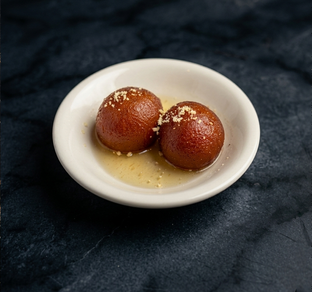 Two dark brown, round gulab jamun pastries in a small white bowl filled with syrup, set against a dark textured surface.