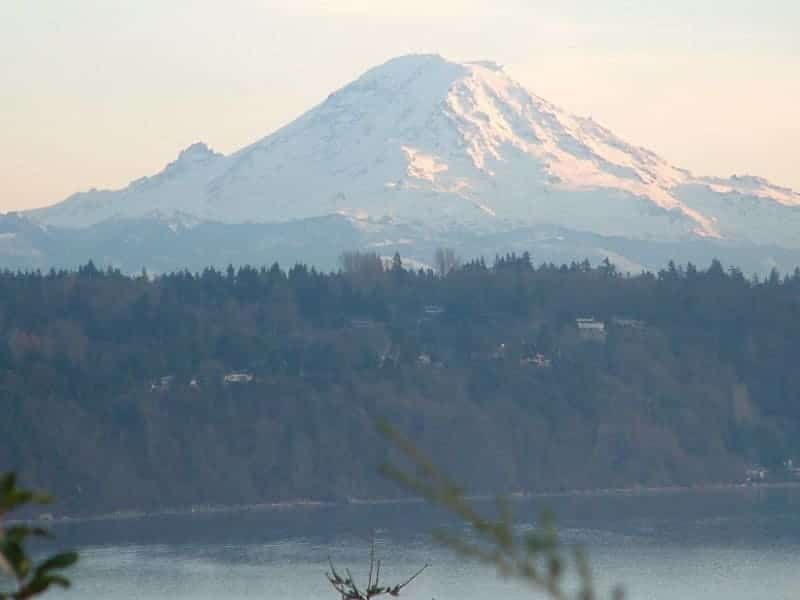 A mountain covered in snow is surrounded by trees and a body of water.
