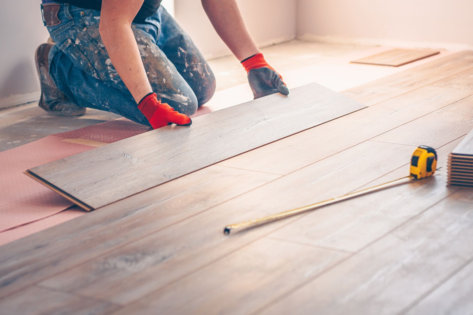 Person installing wooden floorboards, wearing gloves, kneeling in a room. Person installing wooden floorboards, wearing gloves, kneeling in a room.