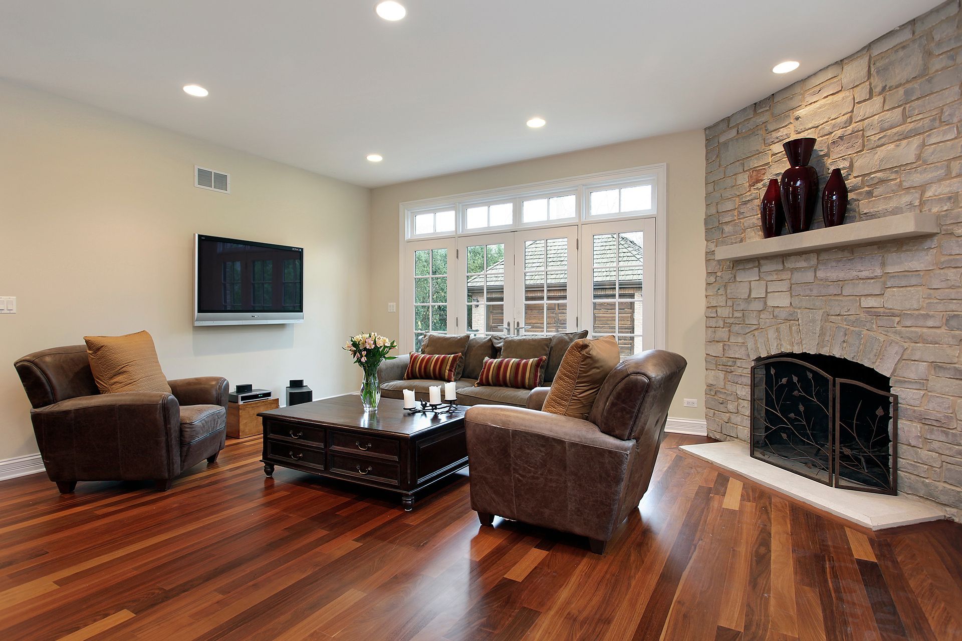 Living room with wood floors, stone fireplace, brown furniture, and large windows. Living room with wood floors, stone fireplace, brown furniture, and large windows.