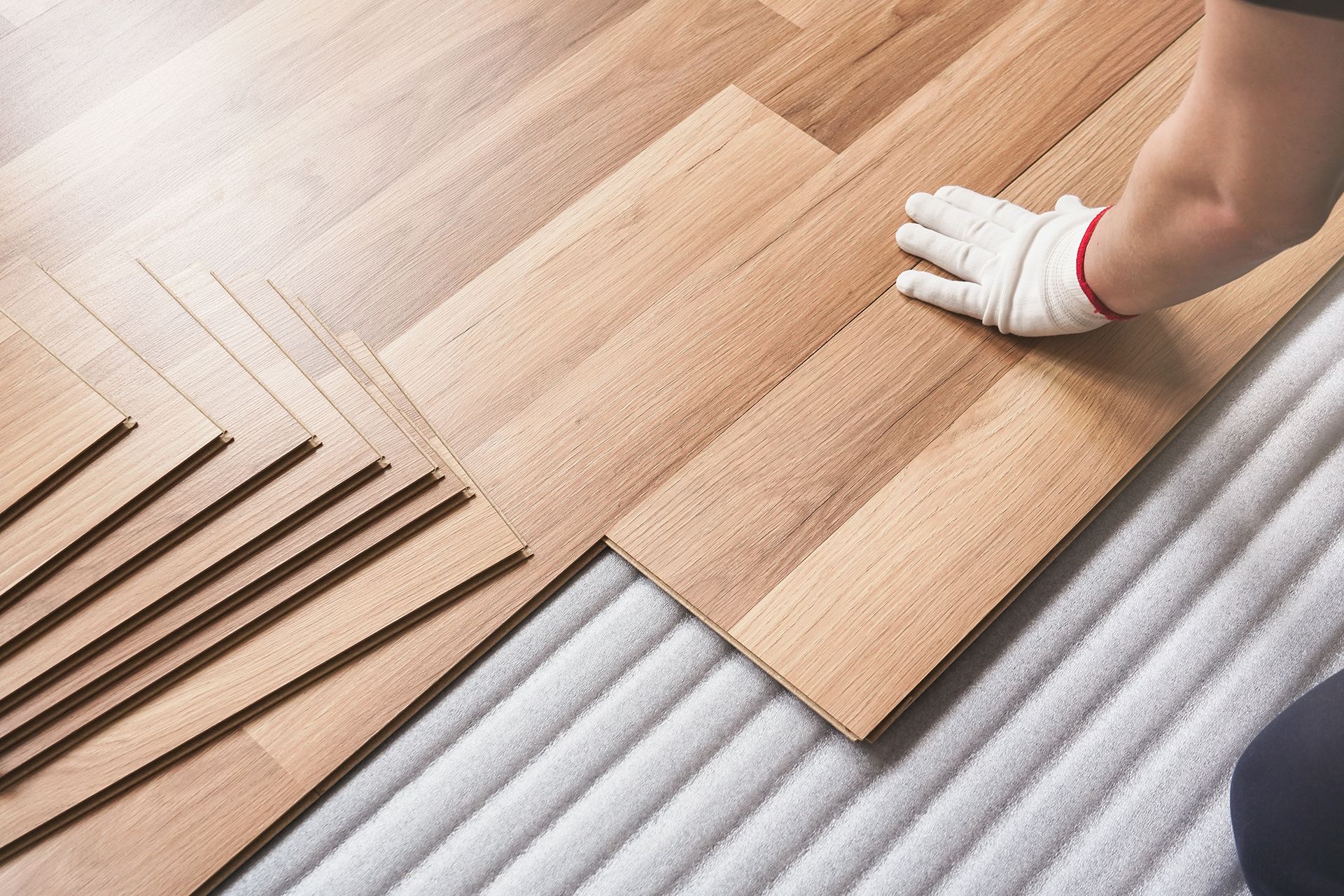 Person wearing gloves installing wooden floorboards on white underlayment. Person wearing gloves installing wooden floorboards on white underlayment.