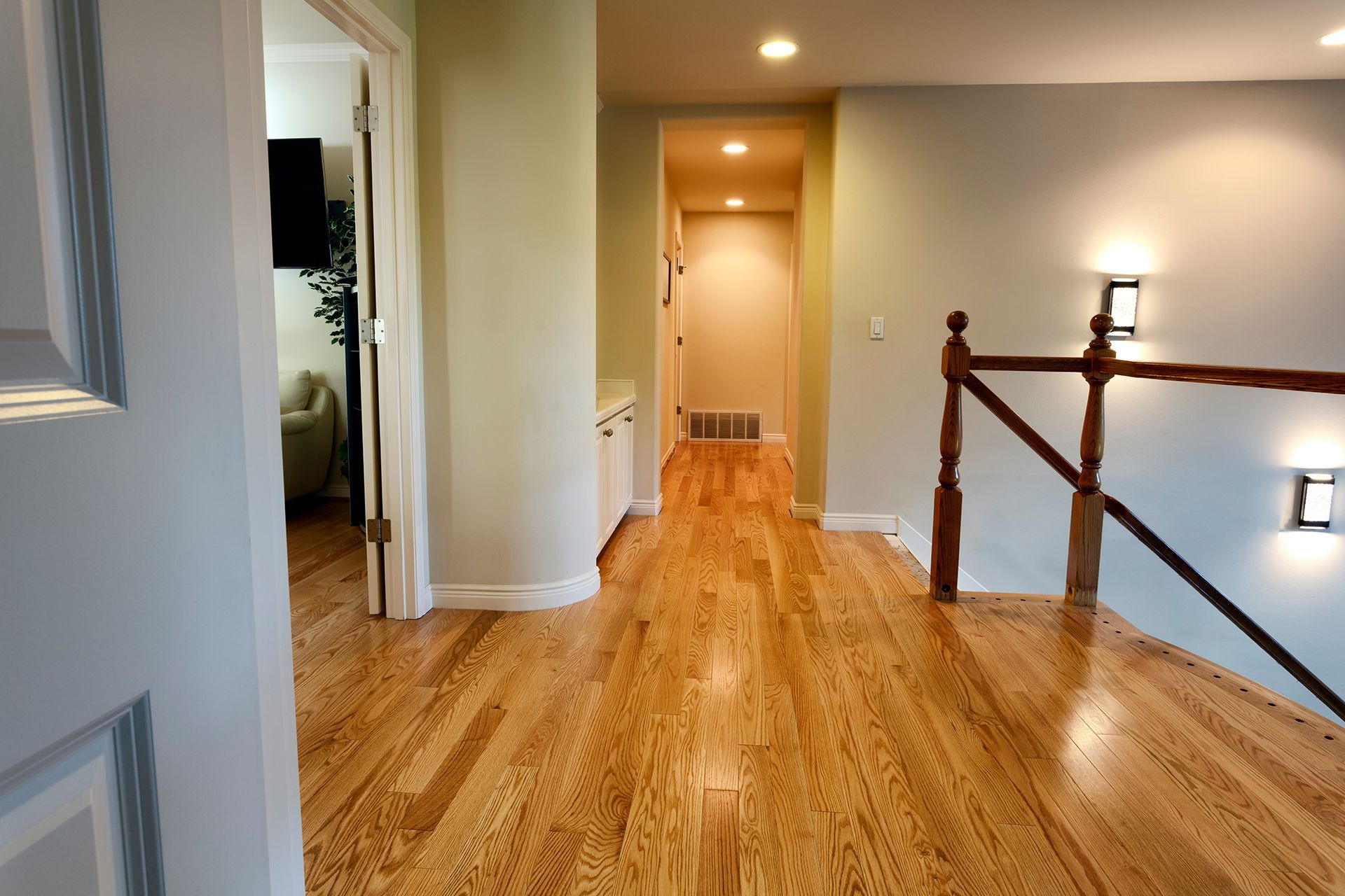 Hallway with hardwood floor, neutral walls, and a staircase railing. Doorway to a living room on the left. Hallway with hardwood floor, neutral walls, and a staircase railing. Doorway to a living room on the left.