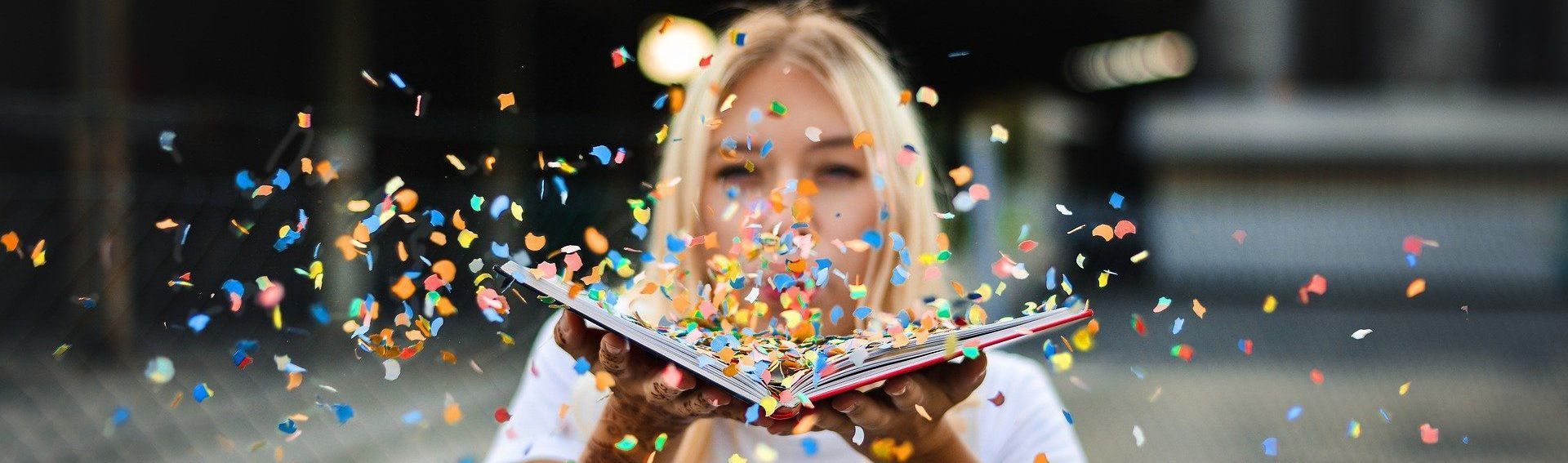 Blonde woman blowing confetti from a book. She is outdoors. The confetti is colorful.