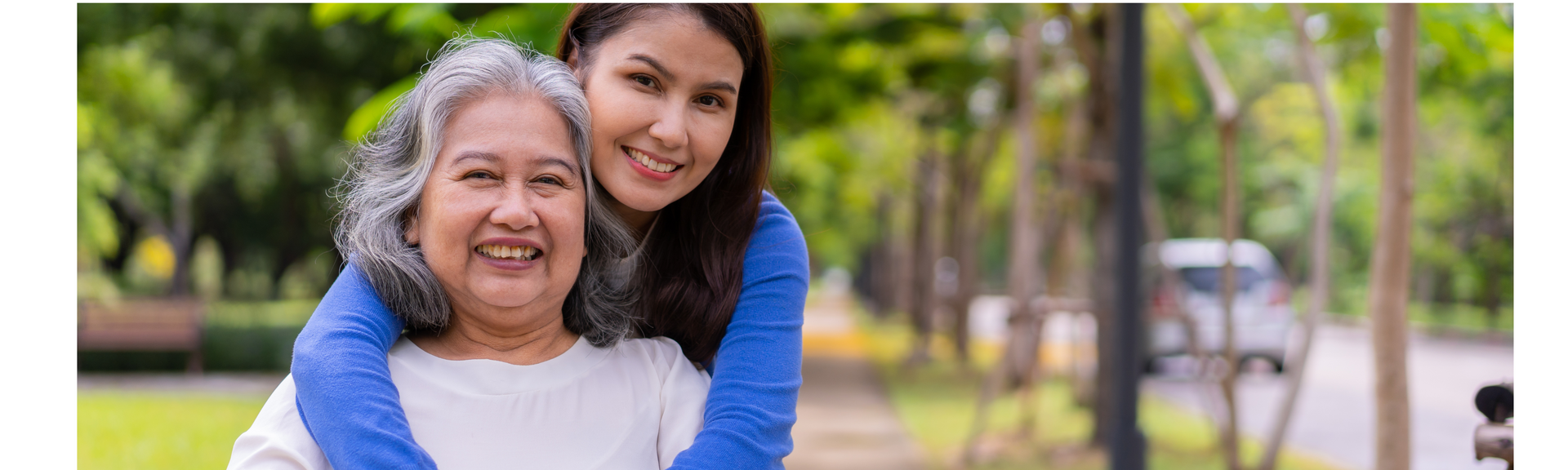 Young woman hugging an older woman, both smiling. Outdoors in a park setting with greenery.
