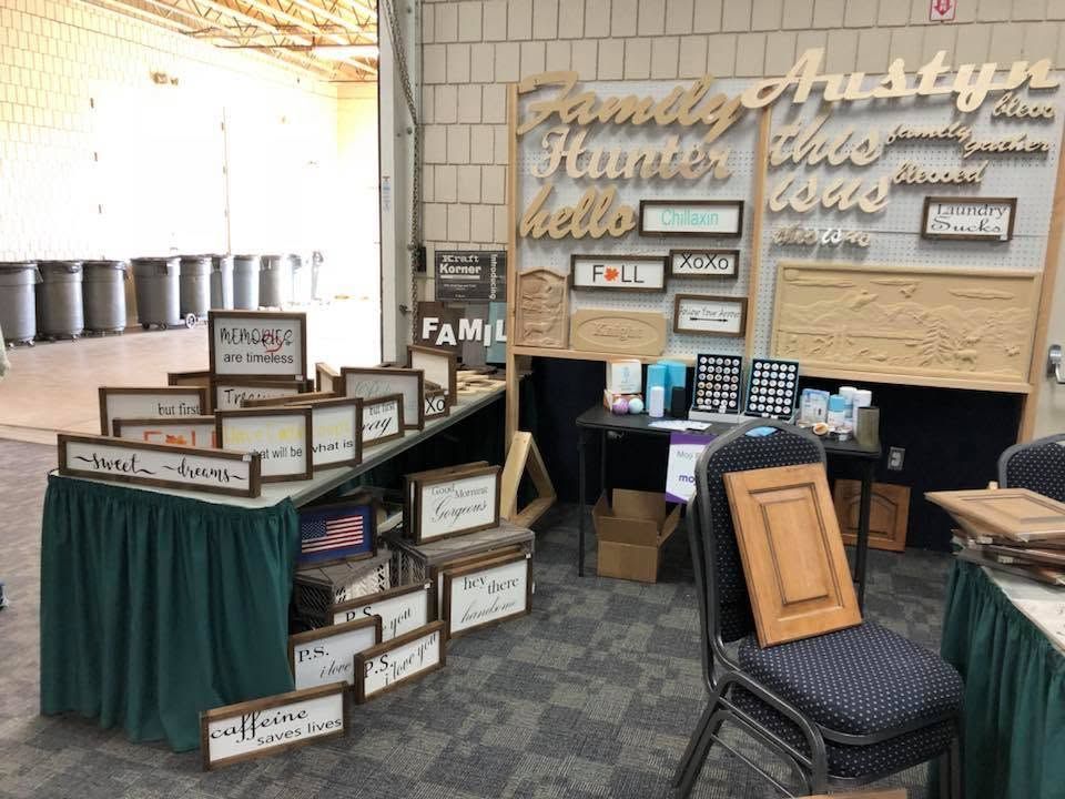 Indoor trade show booth with rustic wooden signs, stacked display boxes, and a table with chairs on carpeted flooring