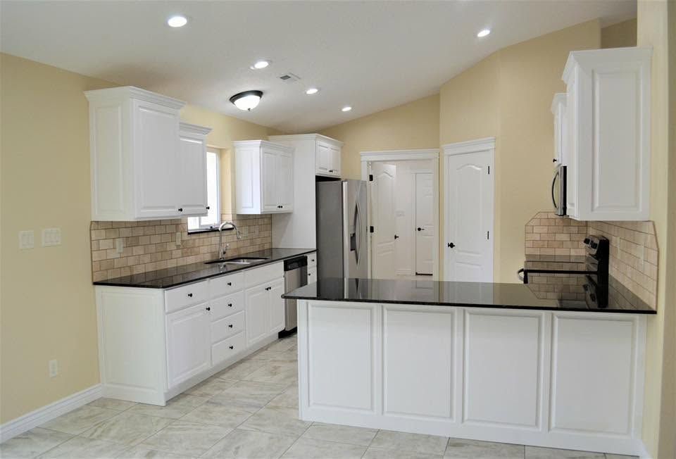 Bright white kitchen with black countertops, stainless refrigerator, and light tile floor