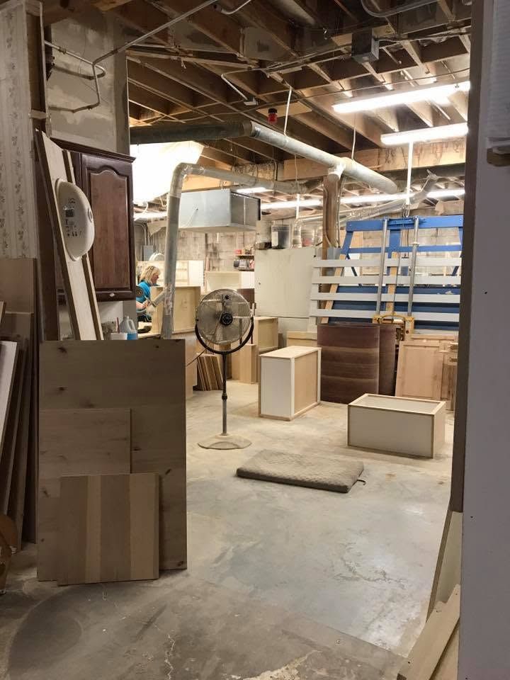 Cluttered workshop with wood cabinets, a standing fan, shelving, and construction materials under exposed beams